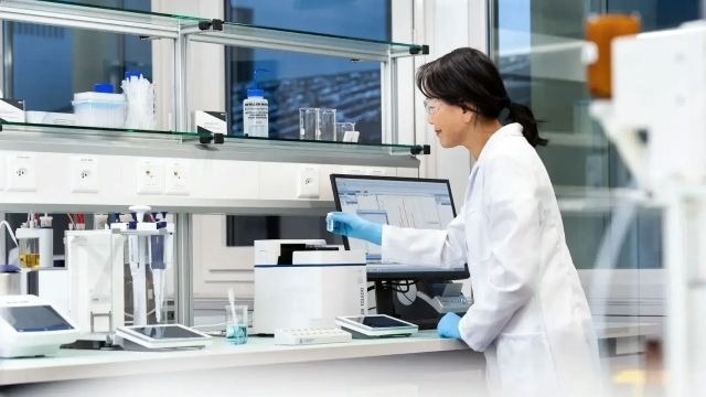 An Ego Pharmaceuticals chemist in the lab. She wears a white labcoat and safety spectacles, with dark hair in a ponytail at the nape of her neck.