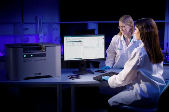Two female scientists wearing lab coats and goggles looking at data on a PC