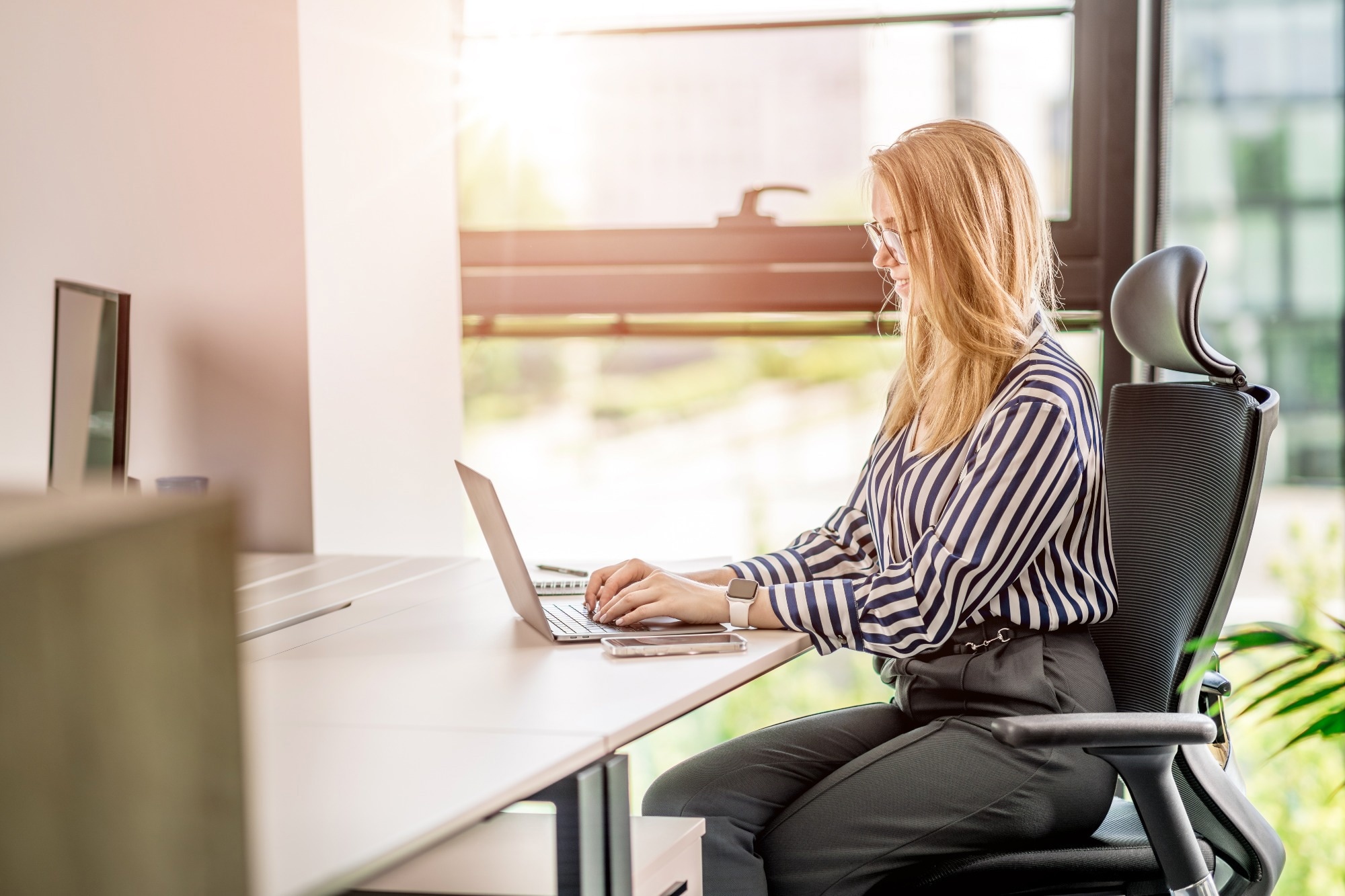 A comfortable place by the window at work. Young woman working on a laptop. Sun rays coming through the window.