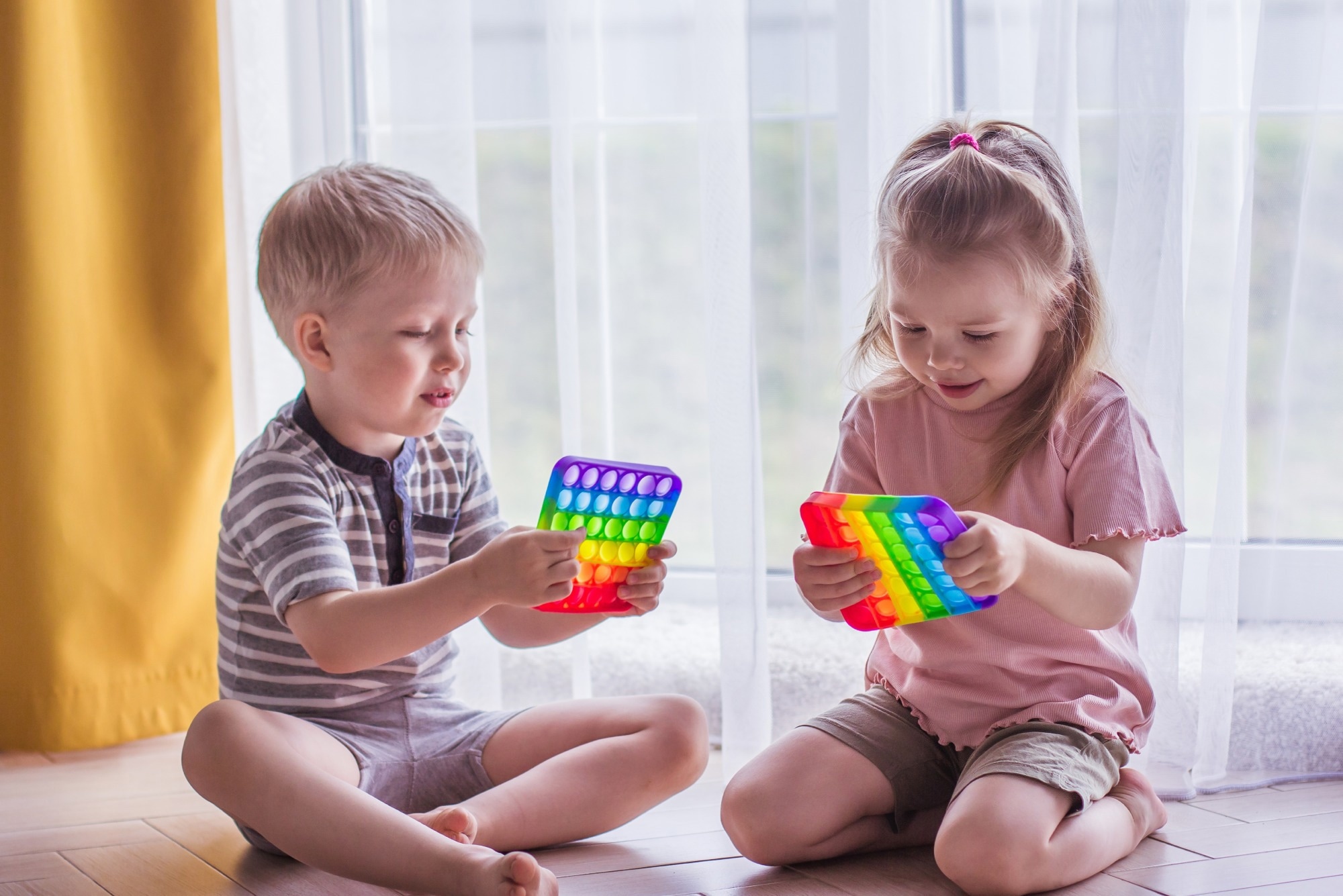 Blonde boy and girl children sit on the floor near the window and play silicone toy antistress pop it. Pop it sensory toy. Stress relief. Colorful anti-stress silicone sensors toy