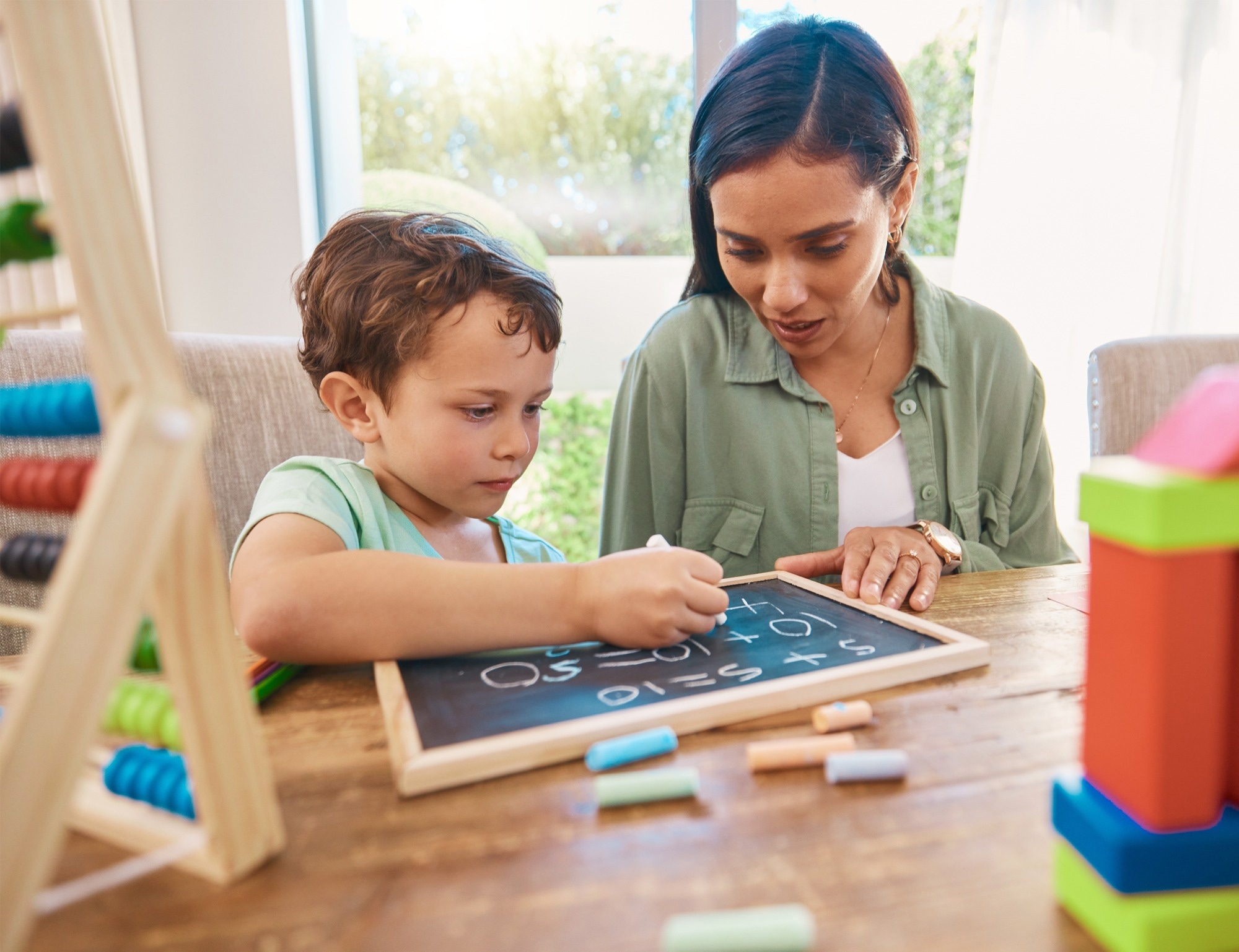 Mother and young child sitting at a table indoors, with the child writing numbers on a small chalkboard while the mother looks on and guides him