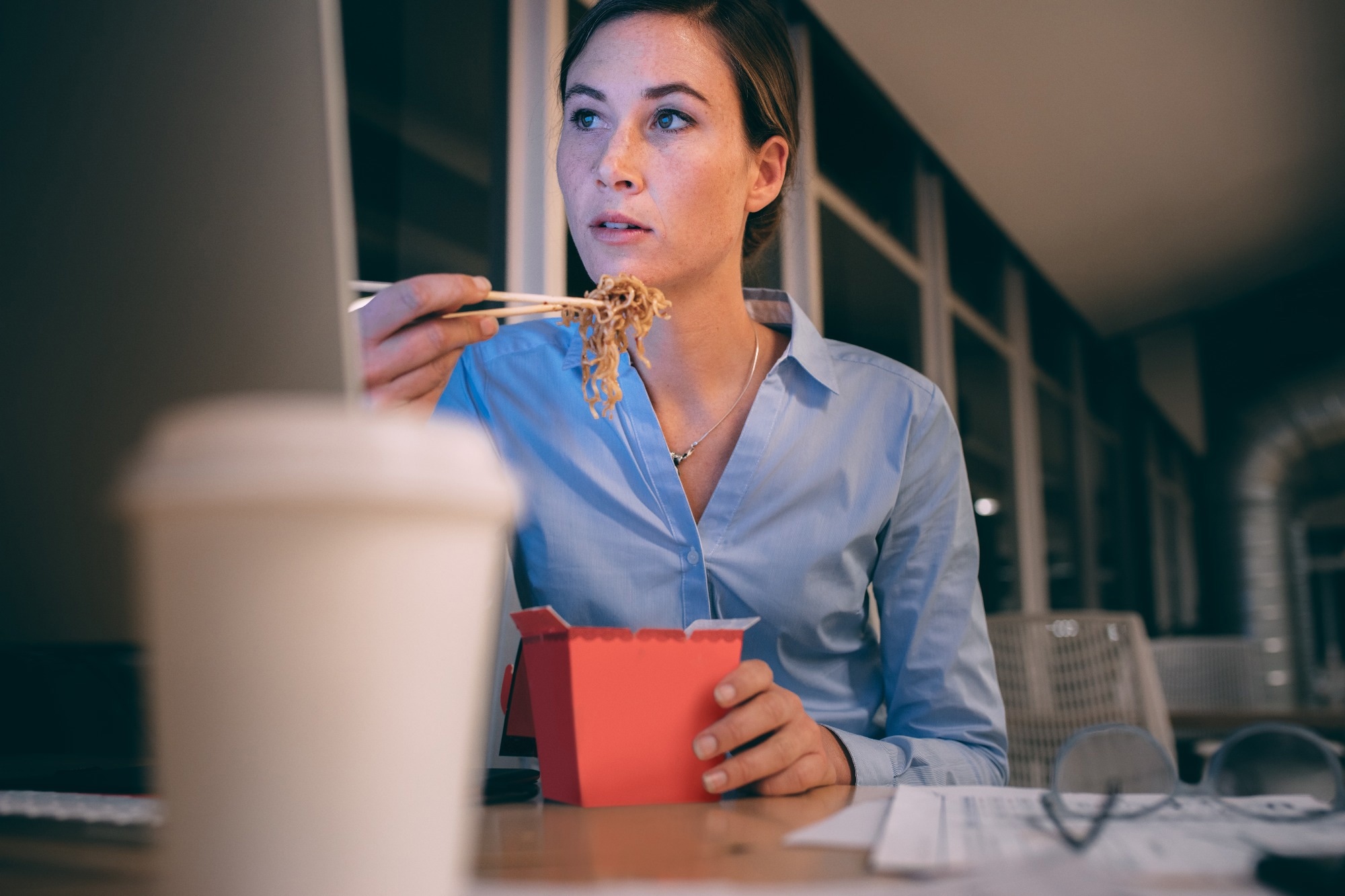 Businesswoman sitting working in office late in the nightre and eating noodles sitting at her desk.