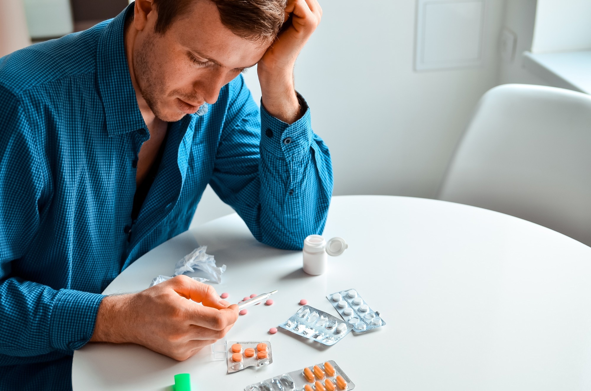 Shot of a young man holding a thermometer and many pills on a table at home. Actual problem, cold and chill, background for medical design