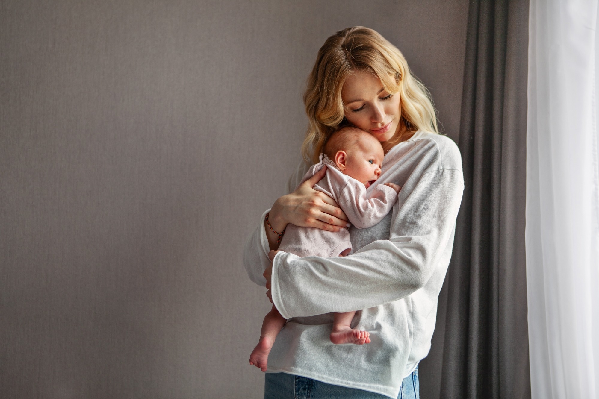 Young mother holds newborn baby while standing at home near window. tender moment with baby dressed in soft pink