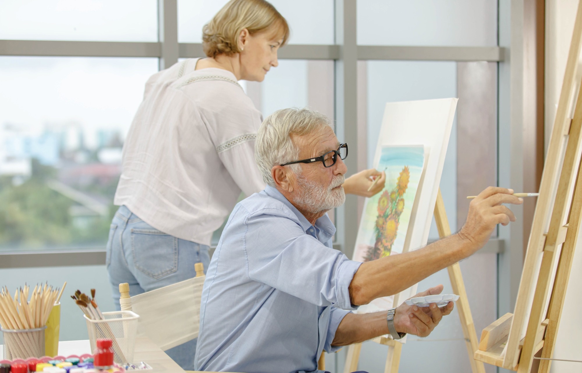 Caucasian old senior elderly male and female artist painter drawing painting flower bouquet picture with watercolor in palette and paintbrush on canvas paper on easel stand together in hobby workshop.
