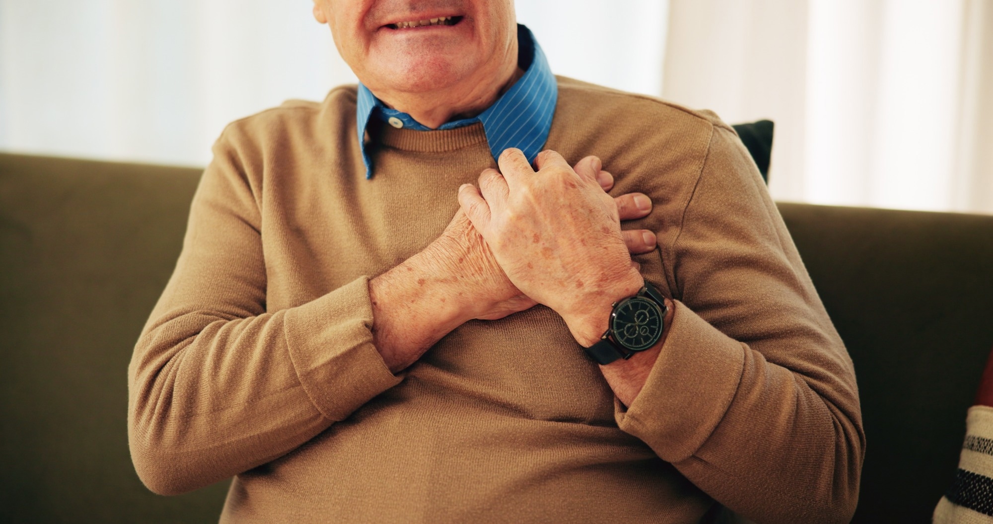 Older man clutching his chest while sitting on a couch, illustrating heart attack symptoms or cardiac chest pain.