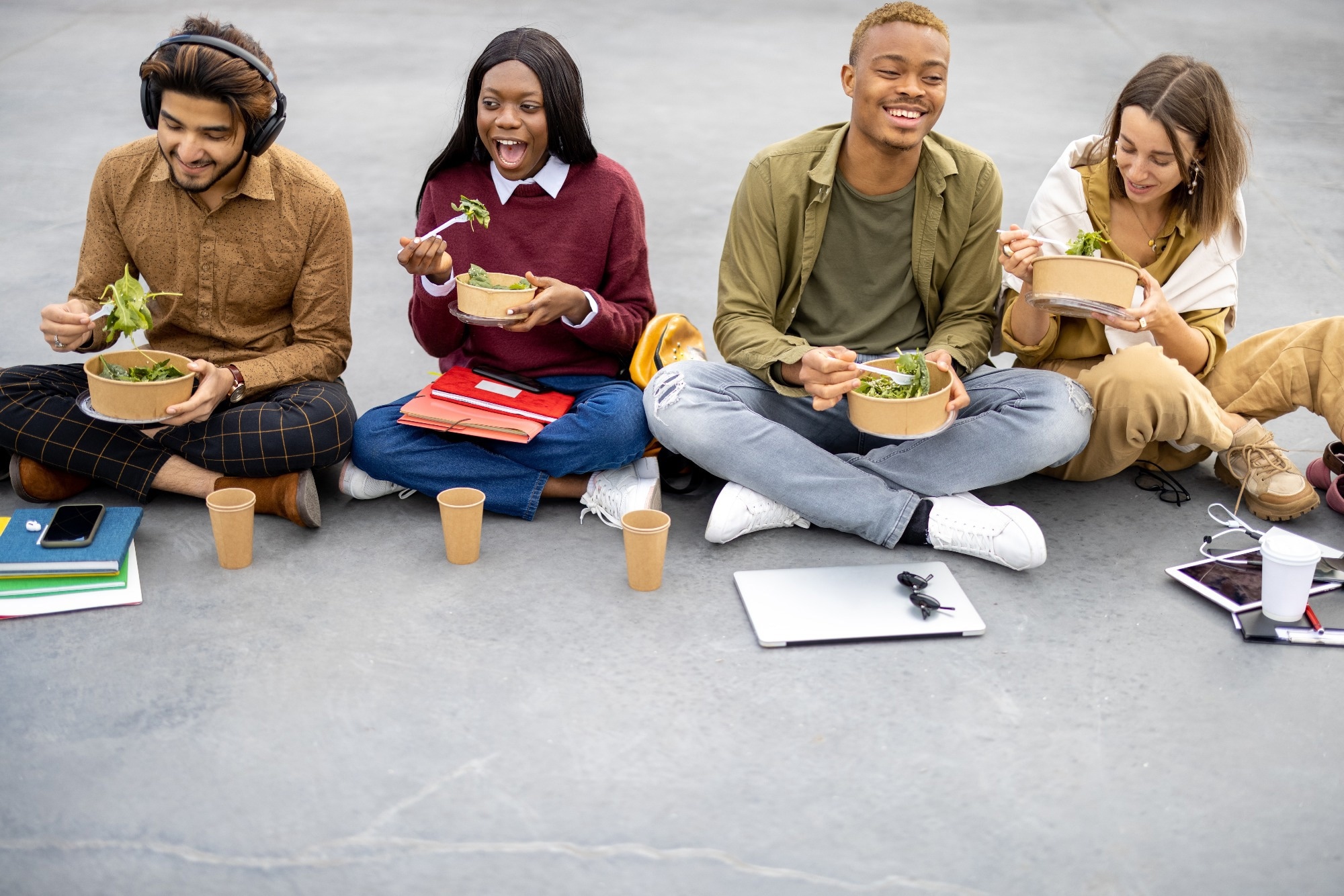 Multiracial students sitting and eating salad during lunch on asphalt at university campus.