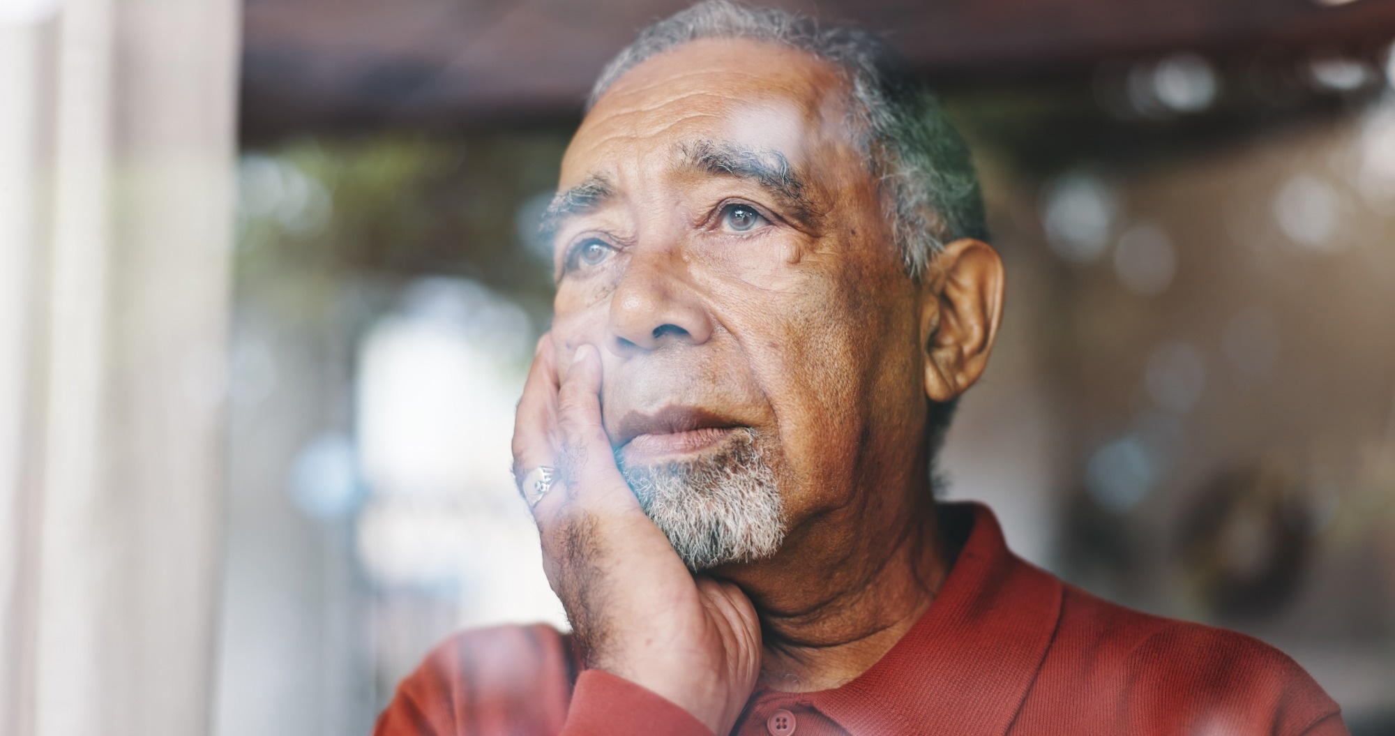 Older black man gazes thoughtfully through a window, resting his face on his hand.