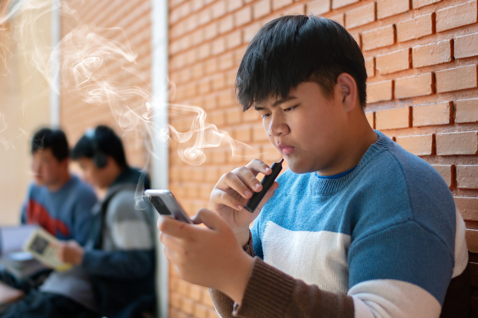 Teen vaping while looking at a smartphone outside a school, highlighting youth e-cigarette use and social media exposure.