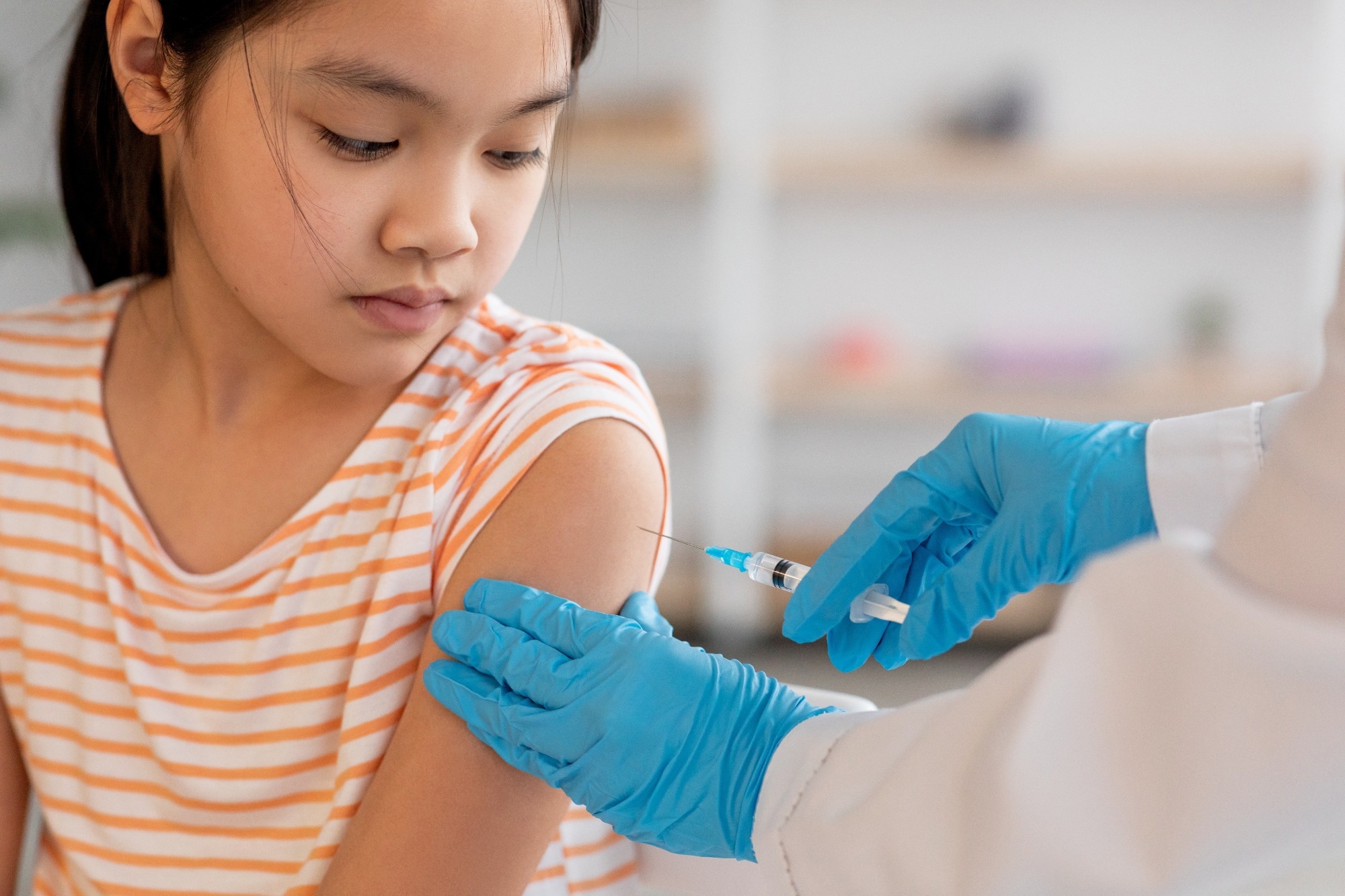 Unrecognizable nurse hands in protective gloves making vaccination against coronavirus of flu for little asian girl, home interior, closeup shot. Kids immunization against various disease concept