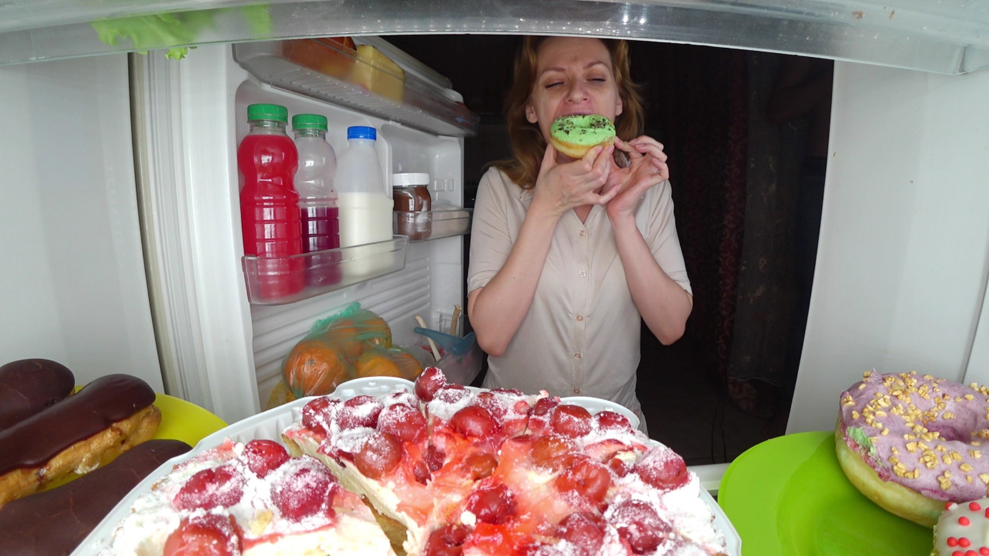 A woman eating a frosted doughnut in front of an open refrigerator filled with desserts and sweet drinks, illustrating emotional eating or binge eating behavior.