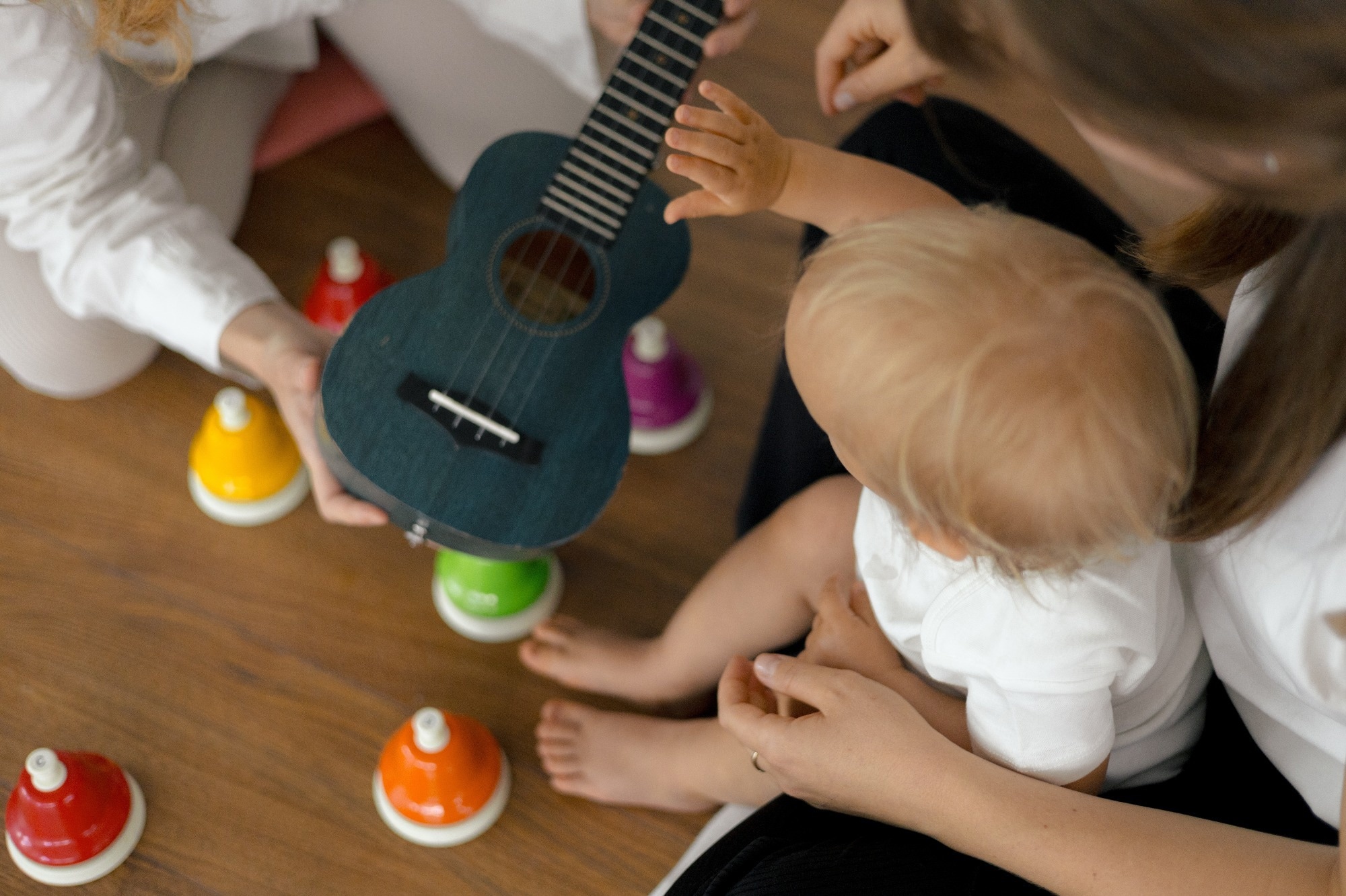 Infant reaches toward a small guitar during a music session with caregivers, illustrating early exposure to musical sounds and interaction.