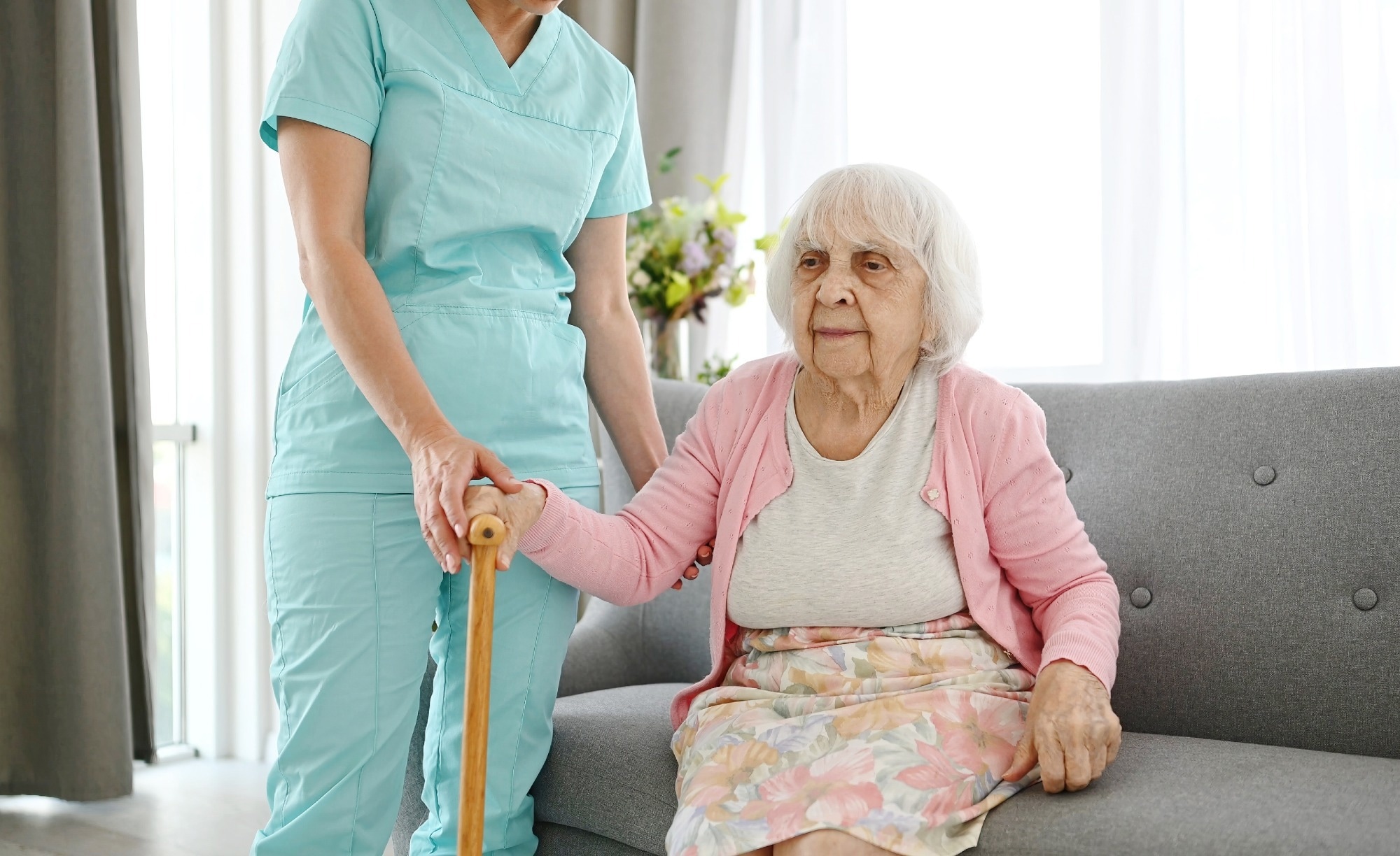 Nurse Hands Cane To Elderly Woman And Helps Her Move Around The Room In A Nursing Home