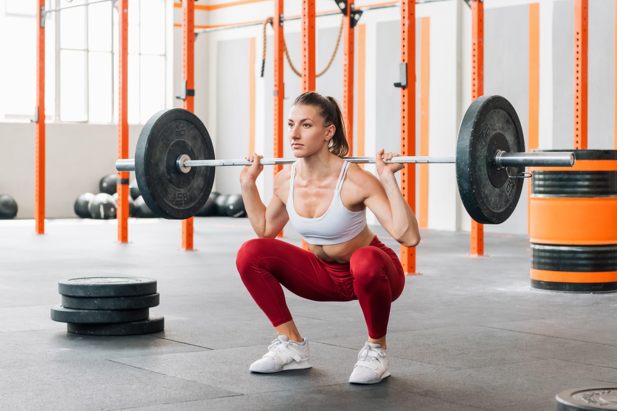 Full body female athlete in activewear looking away and doing barbell back squat during intense workout in sunlit gym