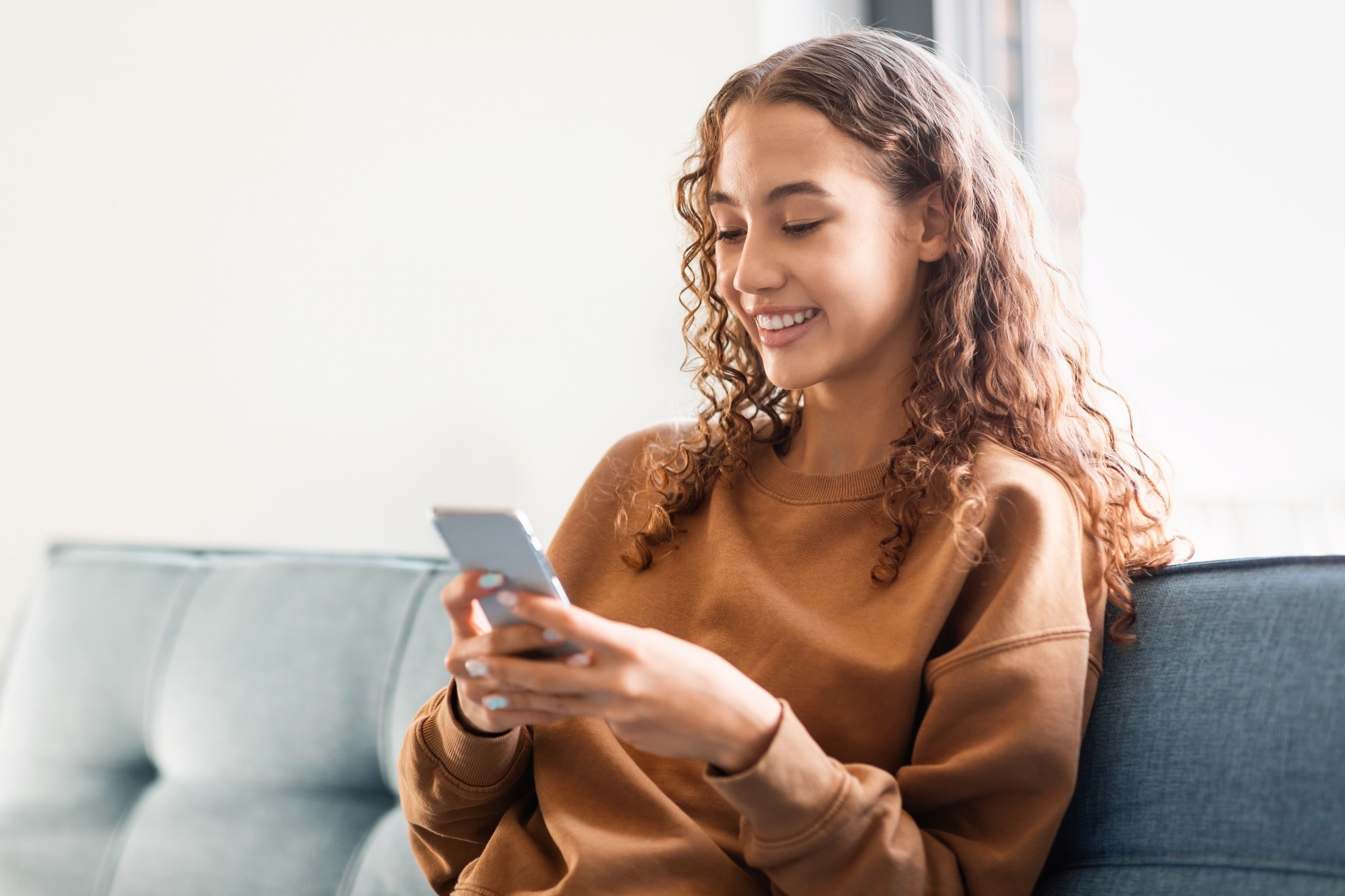 Adolescent girl browsing internet on phone, involved in online communication and application use, sitting carefree and happy in her cozy home setting.