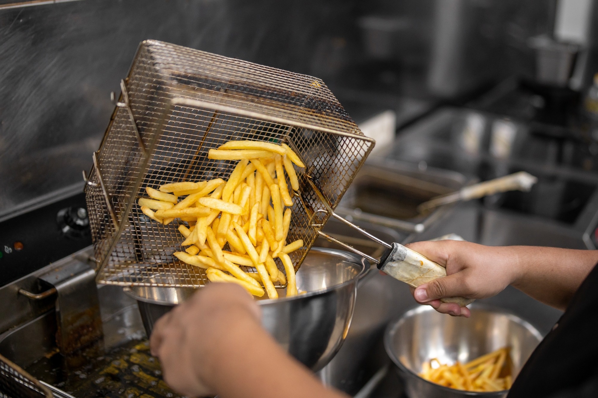 A chef empties fresh, golden french fries from a wire deep fryer basket into a metal bowl in a commercial kitchen.
