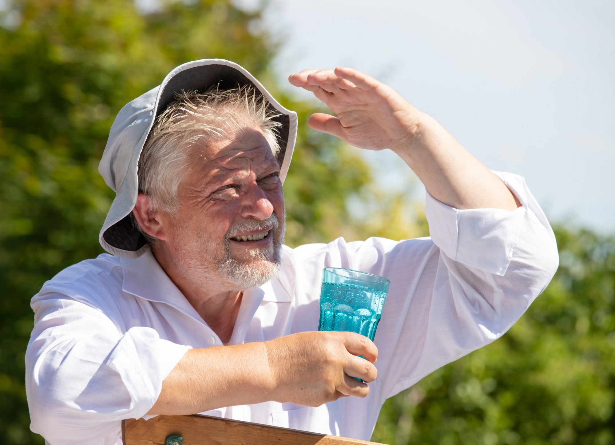 An elderly man in a sun hat sits in the garden in summer and sweats. He drinks water from a glass.